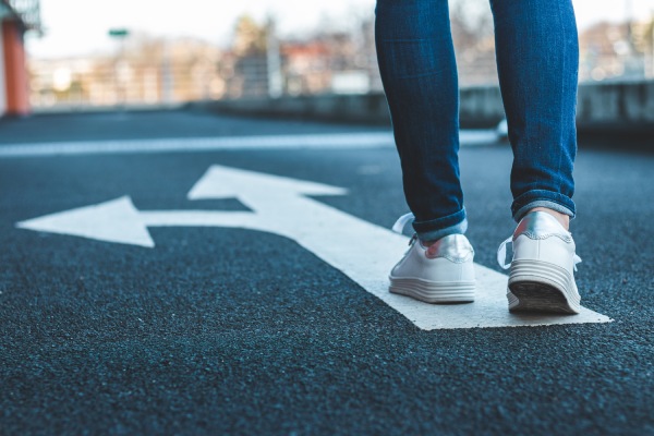 Person in white sneakers and blue jeans standing on a road with a white arrow pointing left and straight.