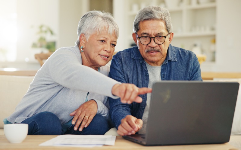 Elderly couple sitting on a couch looking at a laptop while the woman points at the screen.