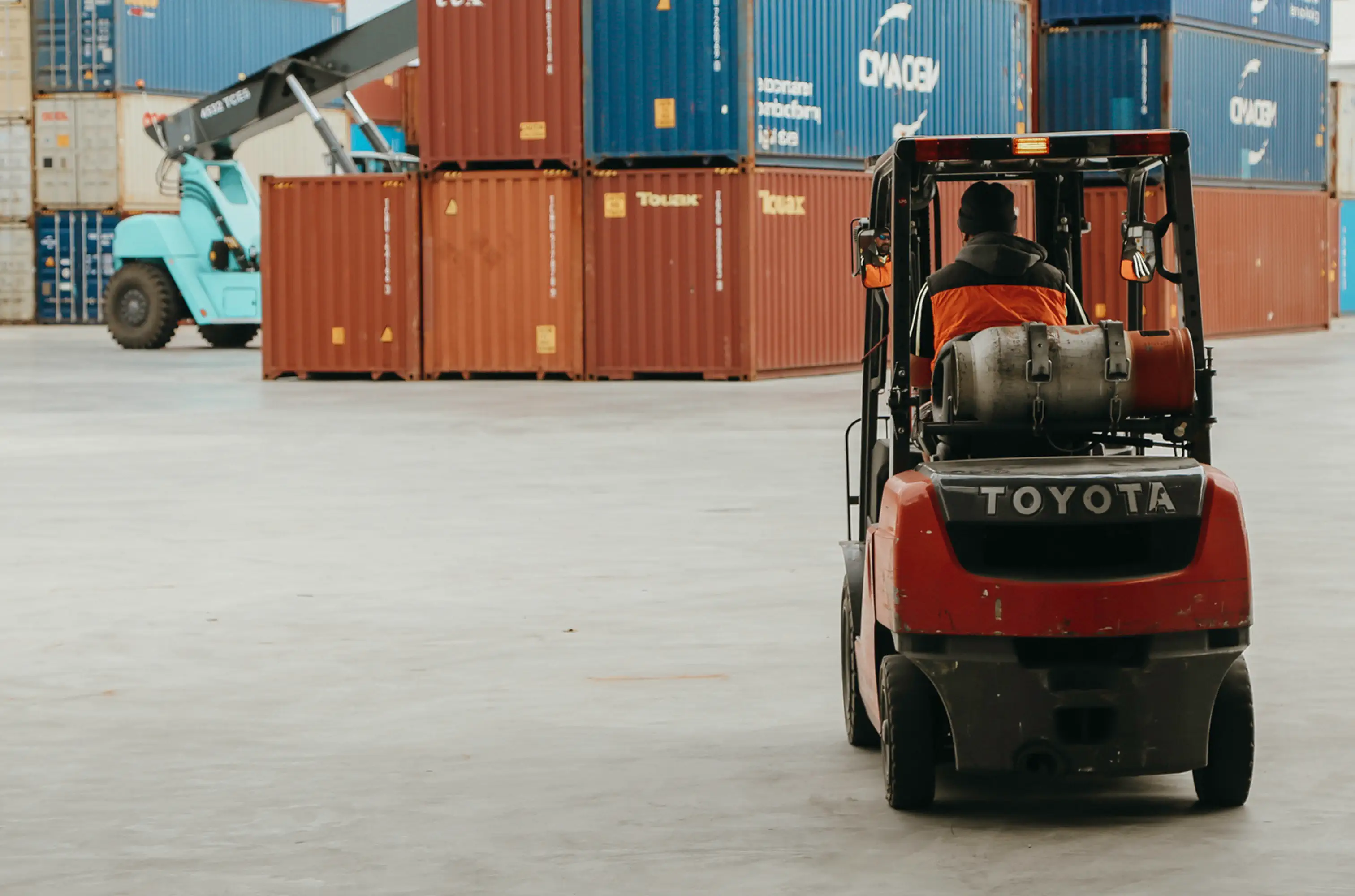 Worker driving a red Toyota forklift in a shipping container yard with stacked containers in the background.