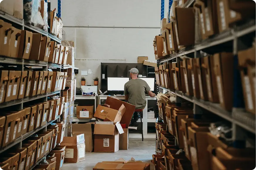 Man working at a desk with computer monitors in a warehouse aisle filled with cardboard boxes on shelves.