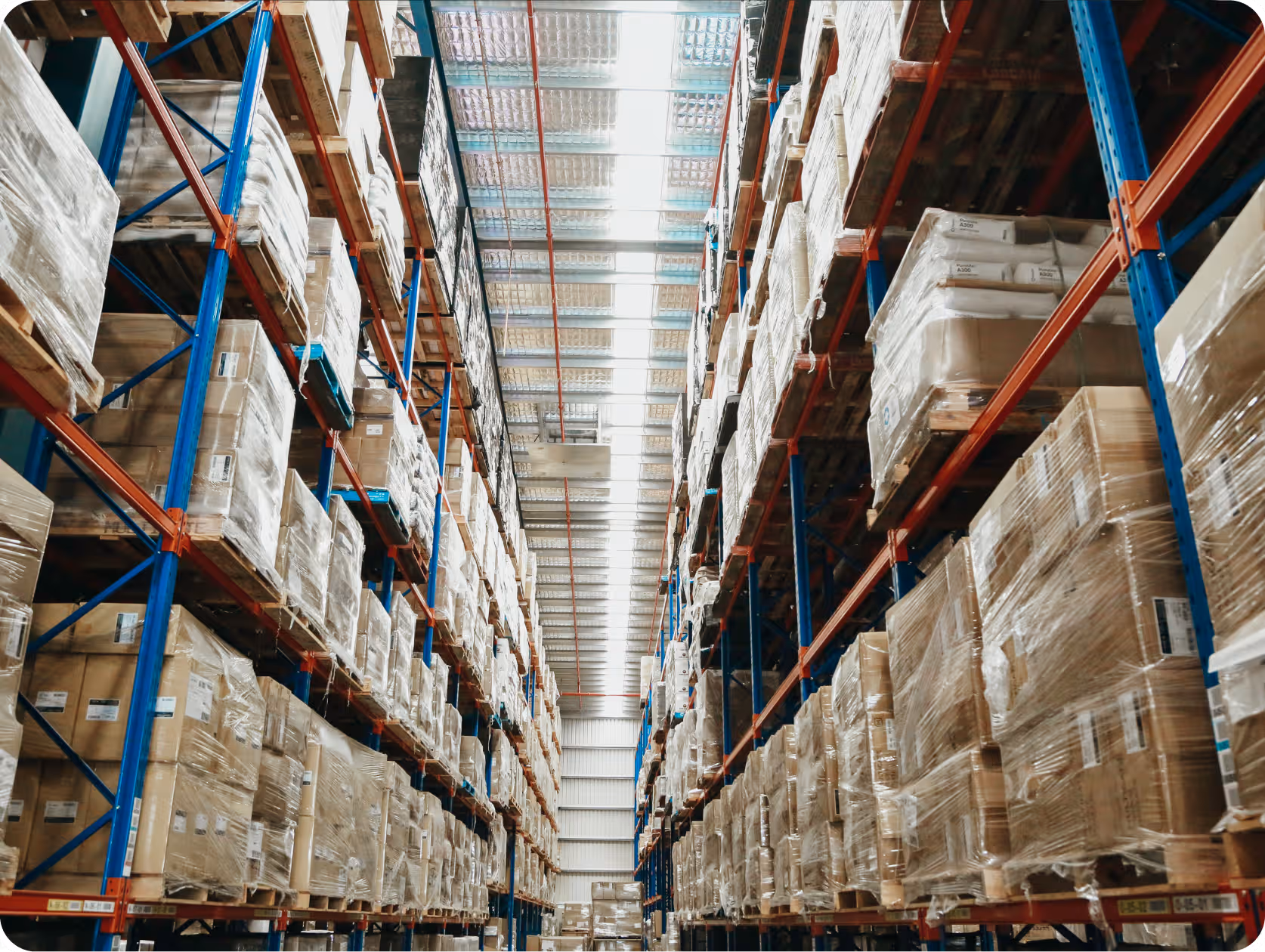 Warehouse aisles with high blue and orange metal shelves fully stacked with plastic-wrapped cardboard boxes.