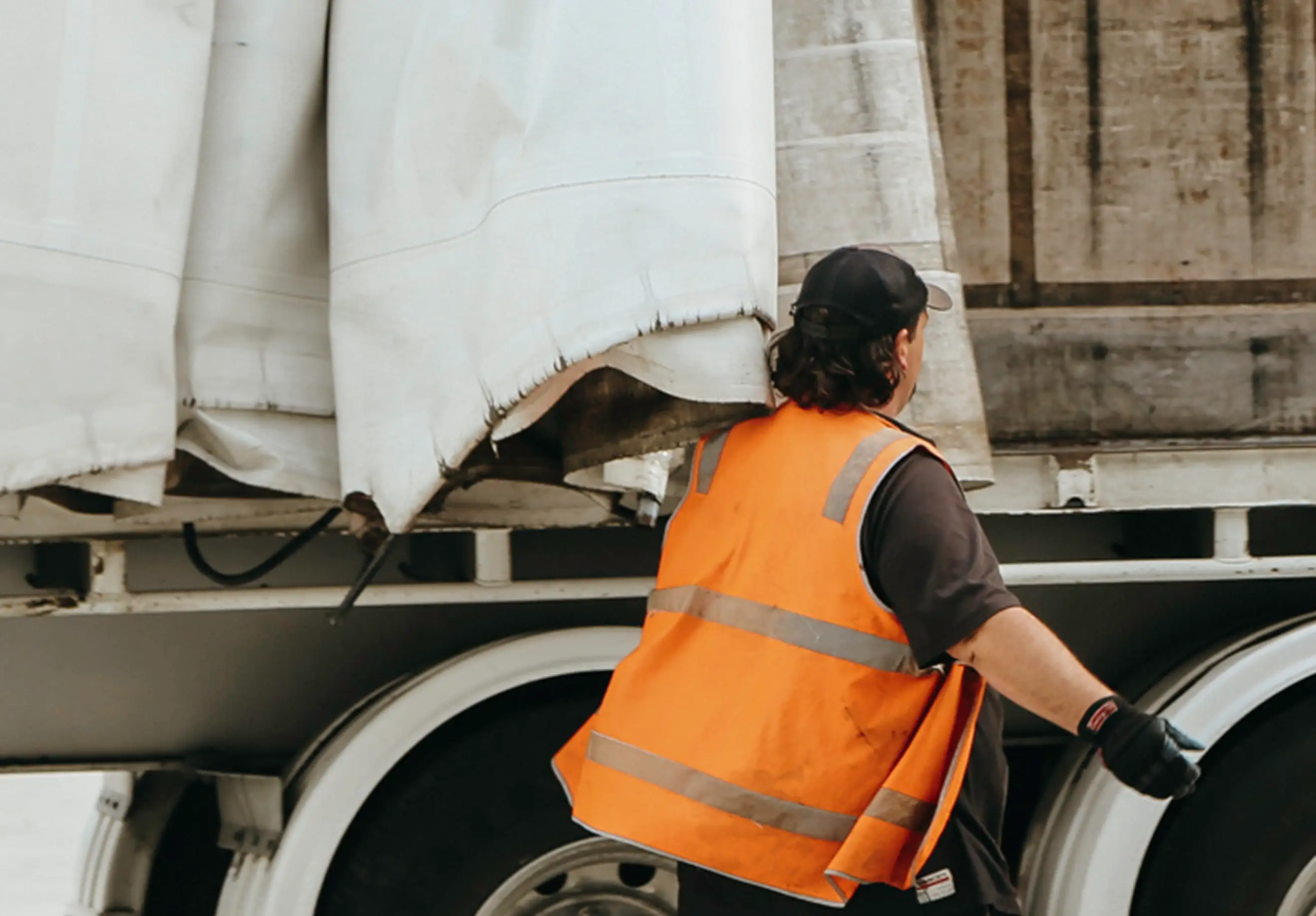 Worker in orange safety vest and black cap standing next to a truck with covered cargo.