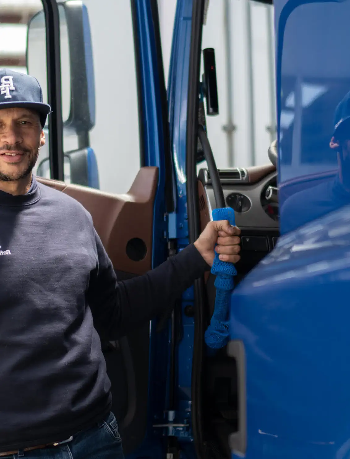 Smiling man wearing a navy cap and shirt, holding onto a blue rope inside a blue truck cabin.