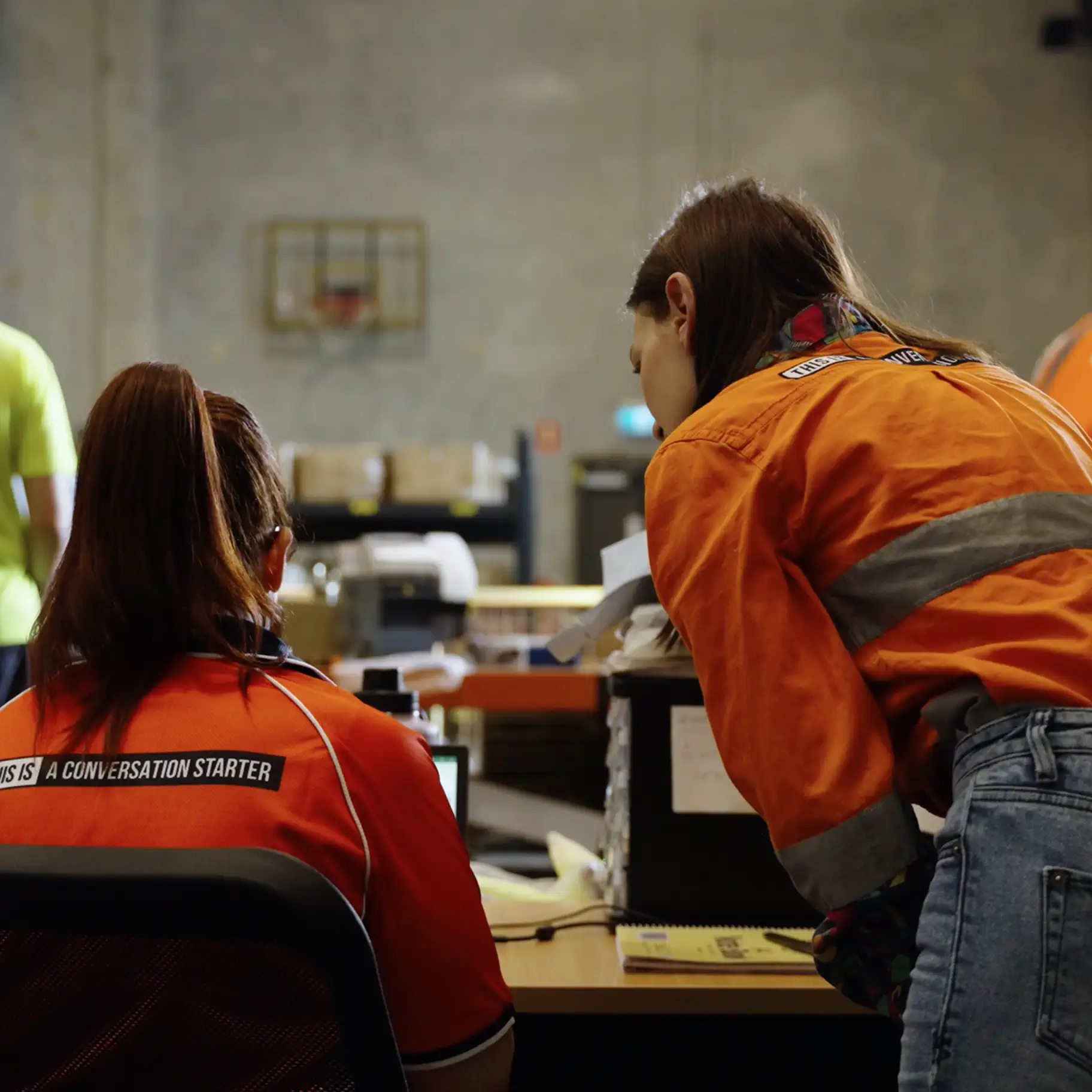 Two people in orange work uniforms collaborating at a desk with industrial shelving in the background.