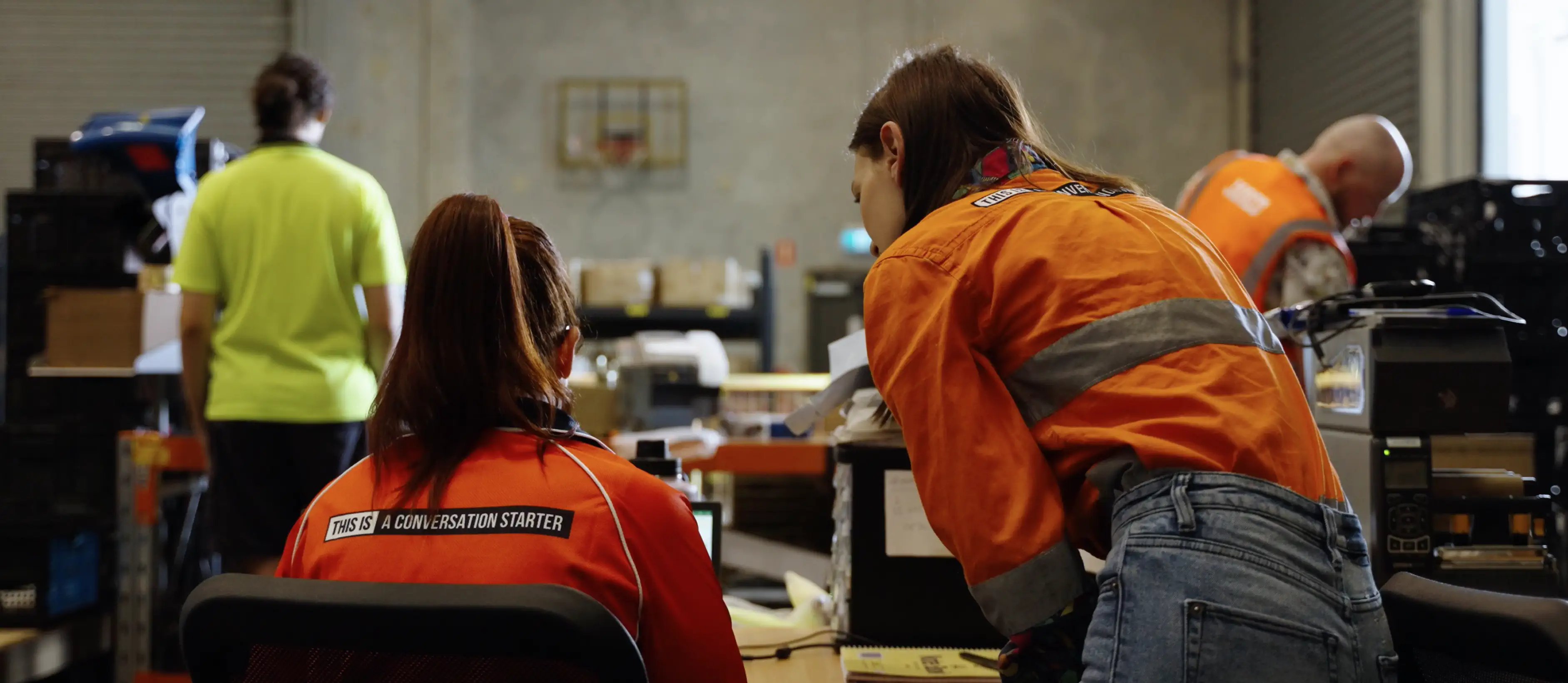 Two workers in high-visibility orange jackets collaborating in an industrial workspace with other workers in the background.