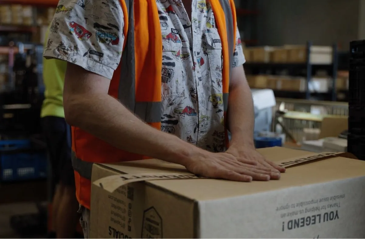 Person wearing an orange safety vest packing a cardboard box in a warehouse.