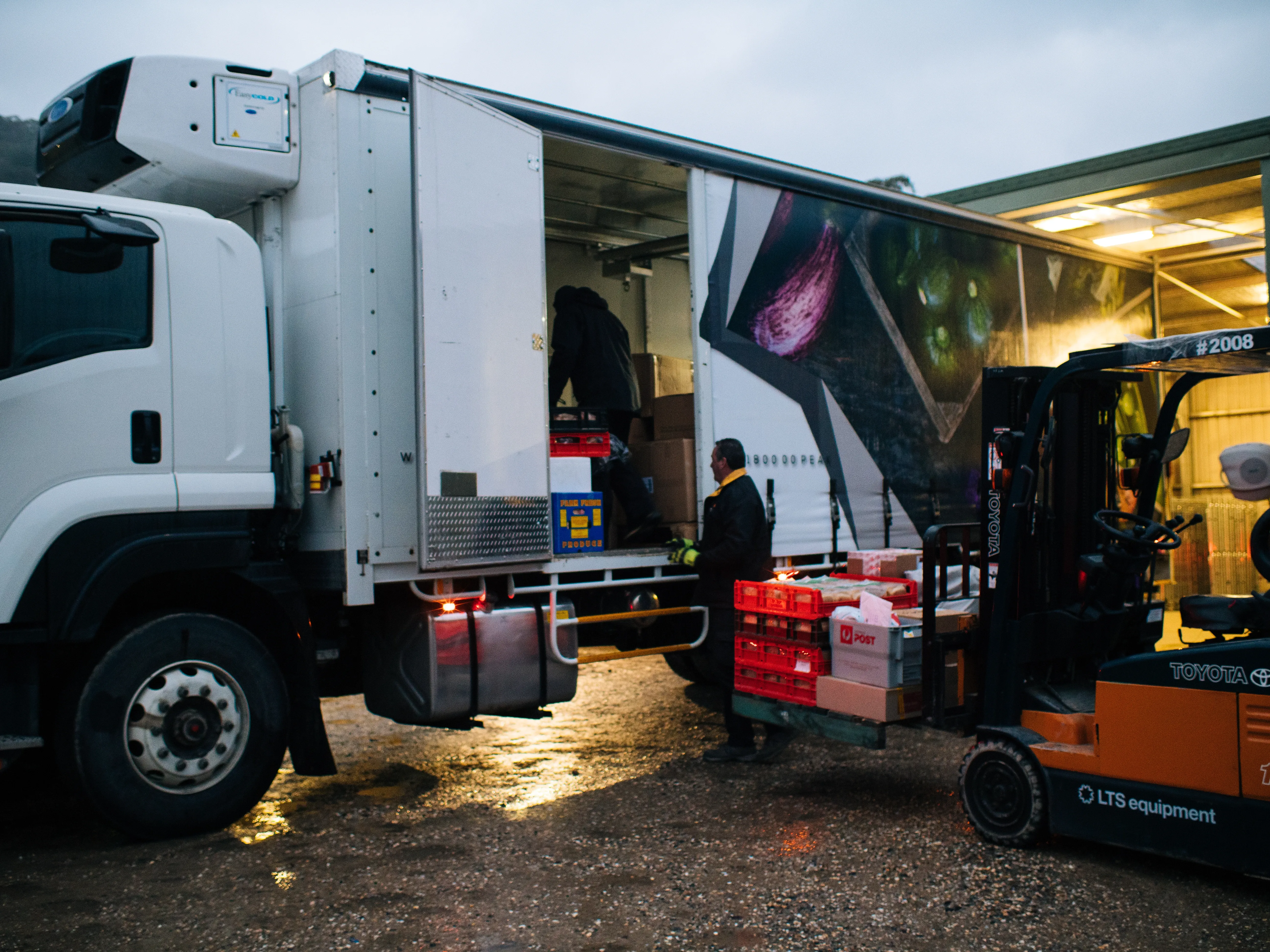 Two workers unloading crates and boxes from a refrigerated delivery truck using a forklift at dusk.