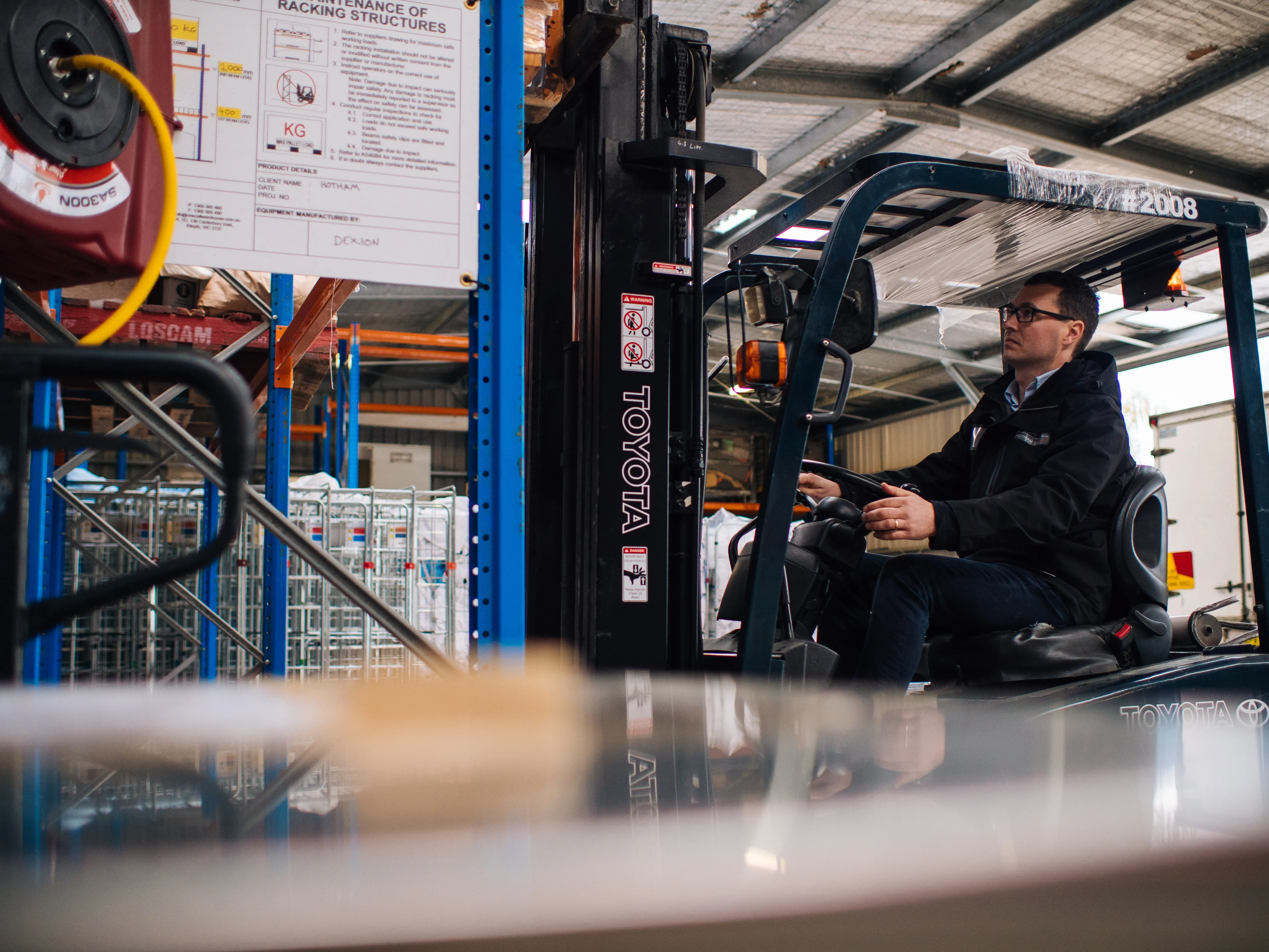 Man in glasses operating a black Toyota forklift inside a warehouse with blue racks.