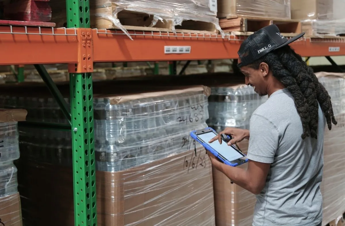 Warehouse worker with long dreadlocks and a black cap using a tablet to check inventory near stacked pallets wrapped in plastic.