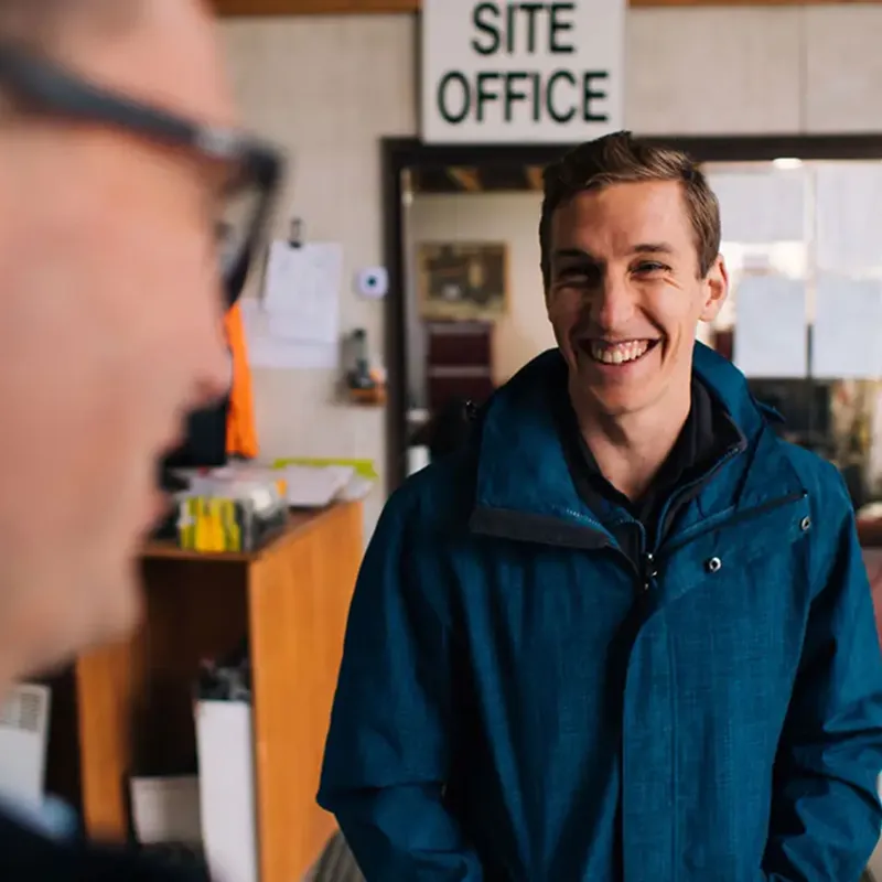 Smiling young man in a blue jacket standing inside an office labeled 'SITE OFFICE' with a blurred person in the foreground.