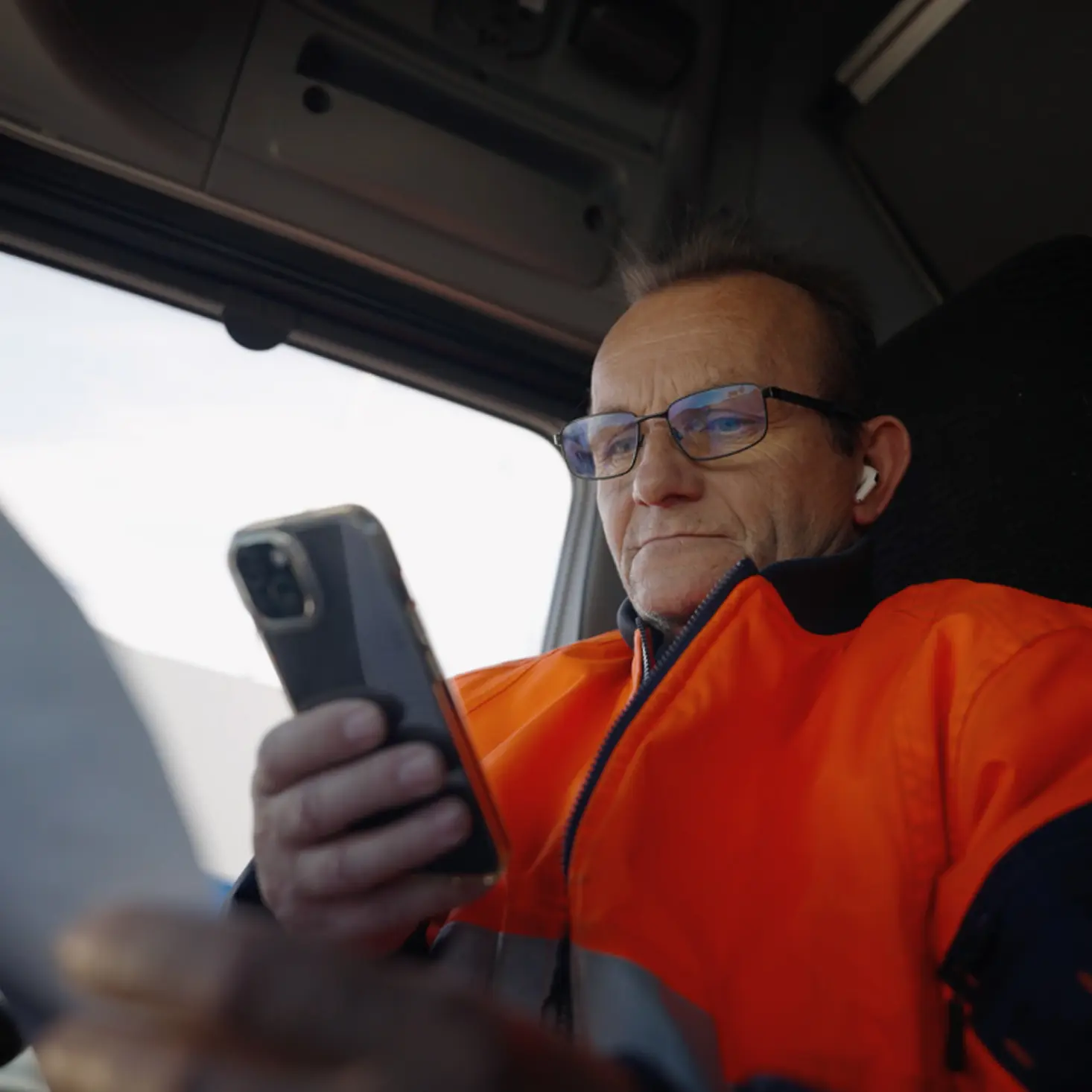 Middle-aged man in orange safety jacket and glasses looking at a smartphone inside a vehicle.