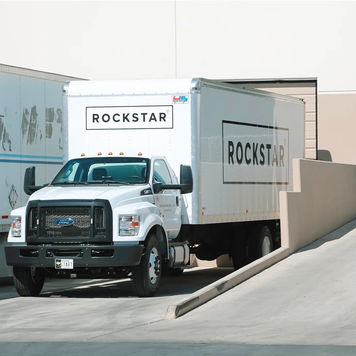 White Ford box truck with ROCKSTAR logo on the side parked near a warehouse loading dock.