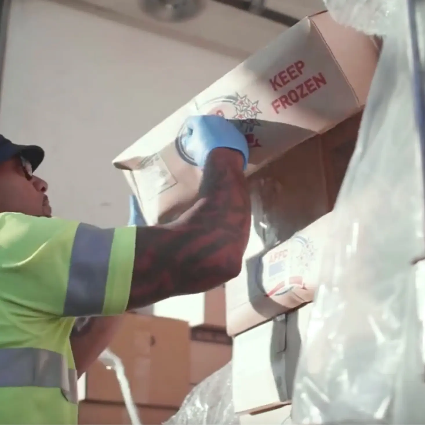 Worker in high-visibility clothing and gloves lifting a cardboard box labeled 'KEEP FROZEN' onto a stack of similar boxes.