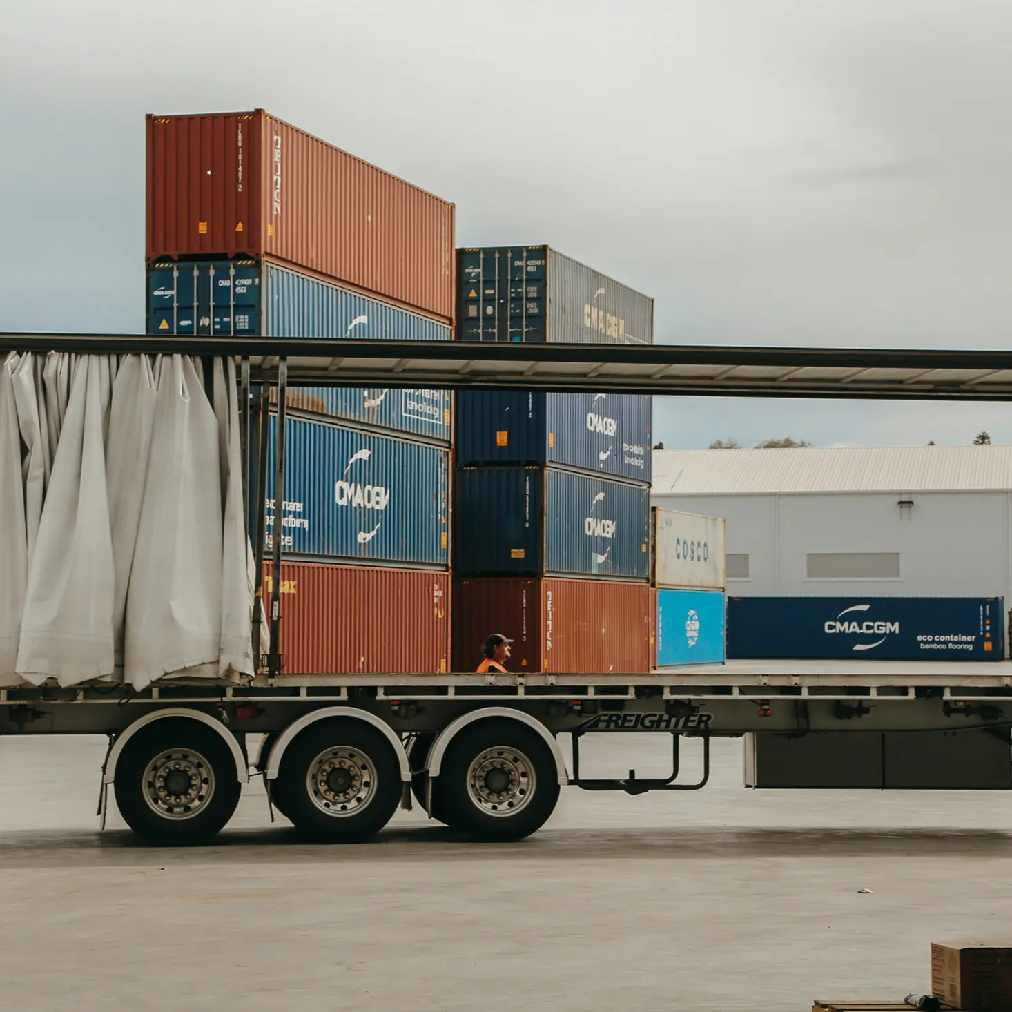 A worker walking beside stacked shipping containers behind a curtain-opened flatbed trailer truck in an industrial yard.