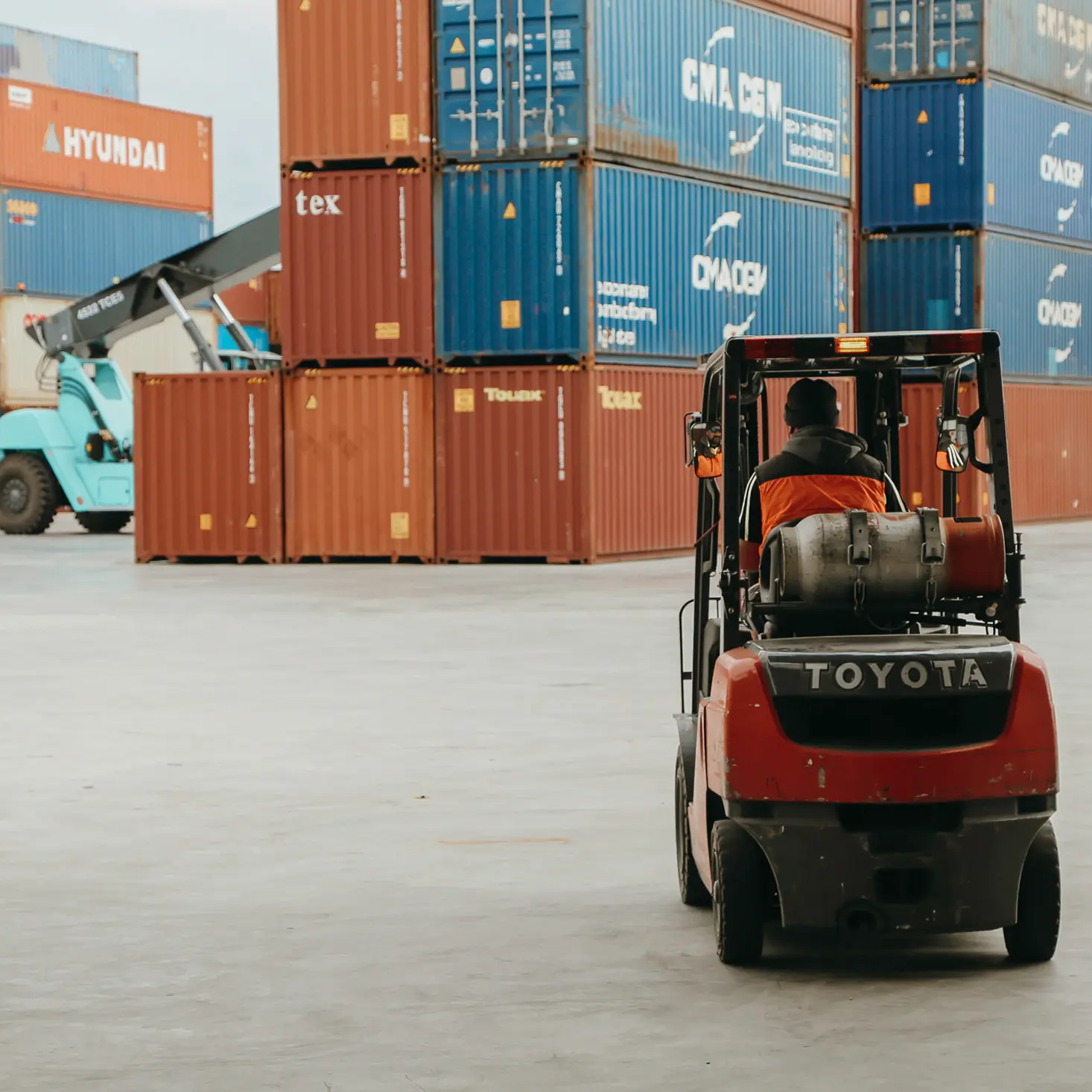 Worker driving a red Toyota forklift in a container yard with stacked shipping containers.