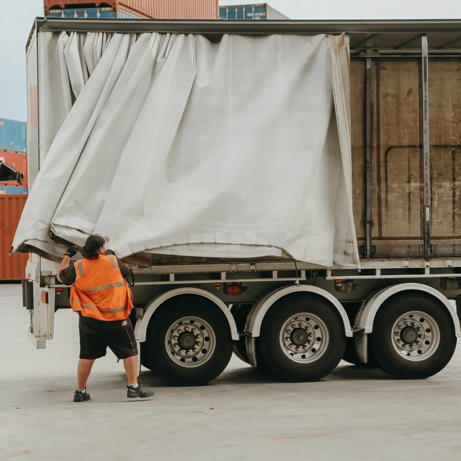 Worker in an orange safety vest pulling back the tarp of a semi-trailer truck at a shipping yard.