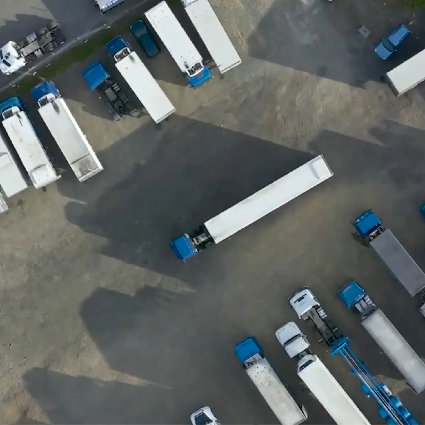 Aerial view of multiple blue trucks with white trailers parked on a dirt lot.