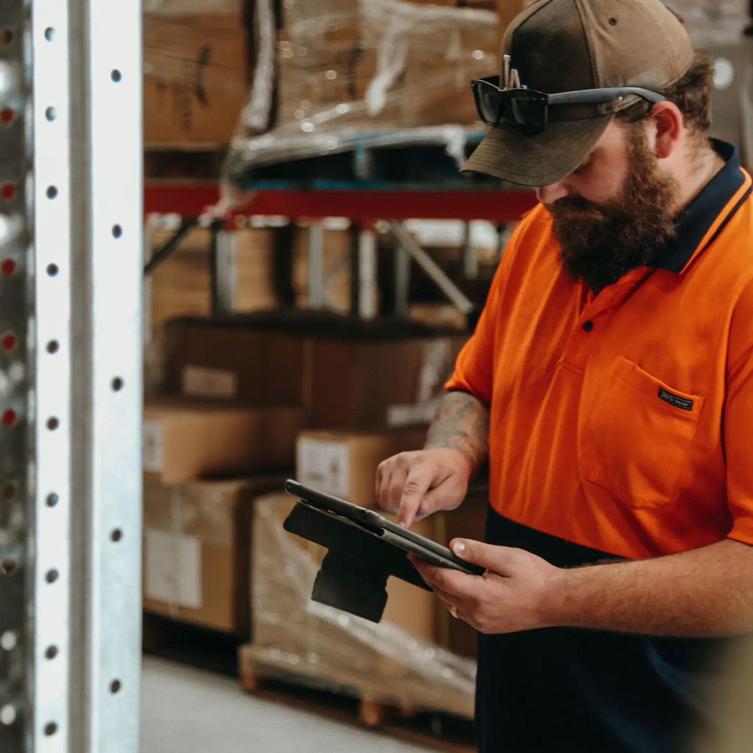 Man in orange and navy work shirt using a tablet in a warehouse with stacked boxes.
