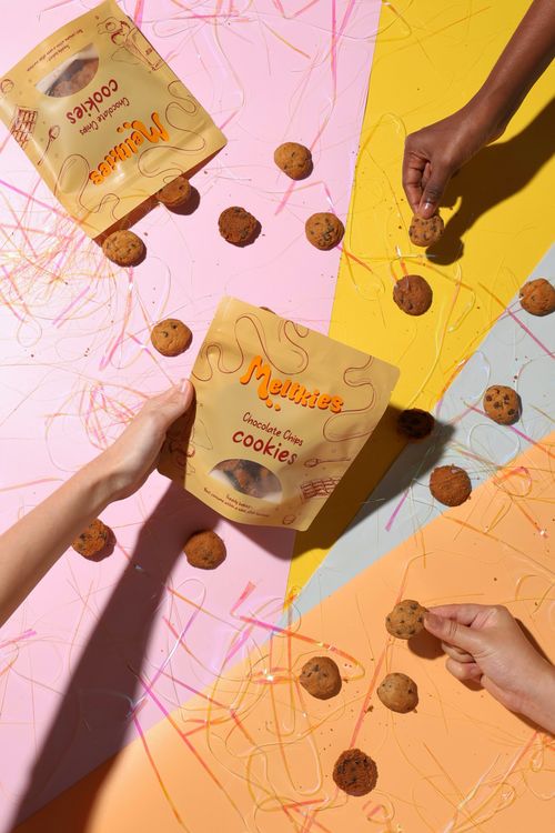 Hands holding Melikies Chocolate Chips Cookies packets with chocolate chip cookies scattered on a colorful pink, yellow, orange, and blue surface.