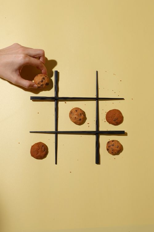 Hand placing a chocolate chip cookie on a black chopstick tic-tac-toe grid with scattered cookie crumbs on a yellow surface.