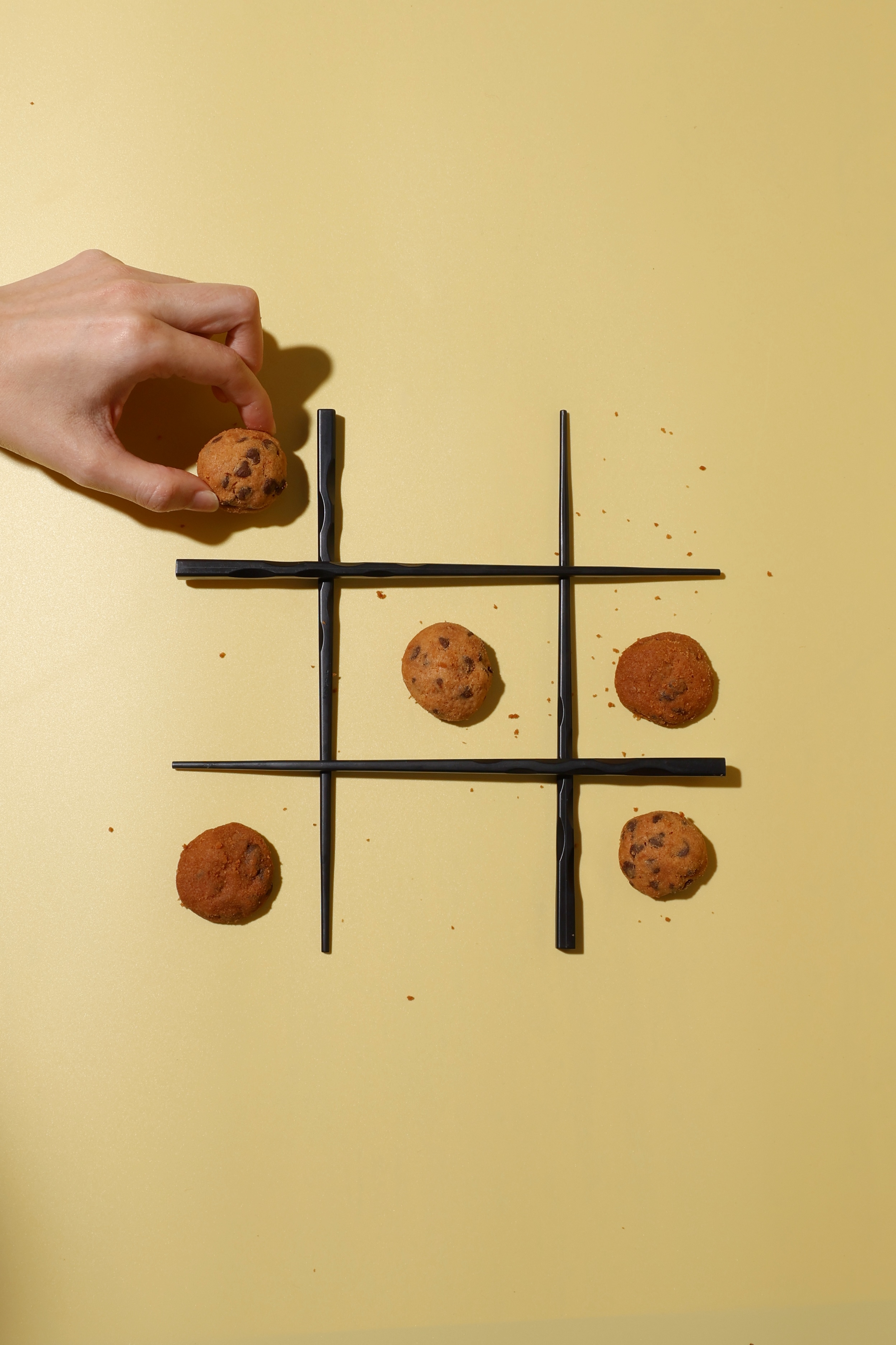 Hand placing a chocolate chip cookie on a black chopstick tic-tac-toe grid with scattered cookie crumbs on a yellow surface.