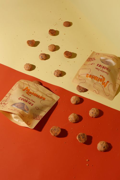 Two Melkkies Chocolate Chips Cookies packages with scattered small round cookies and crumbs on a yellow and red surface.