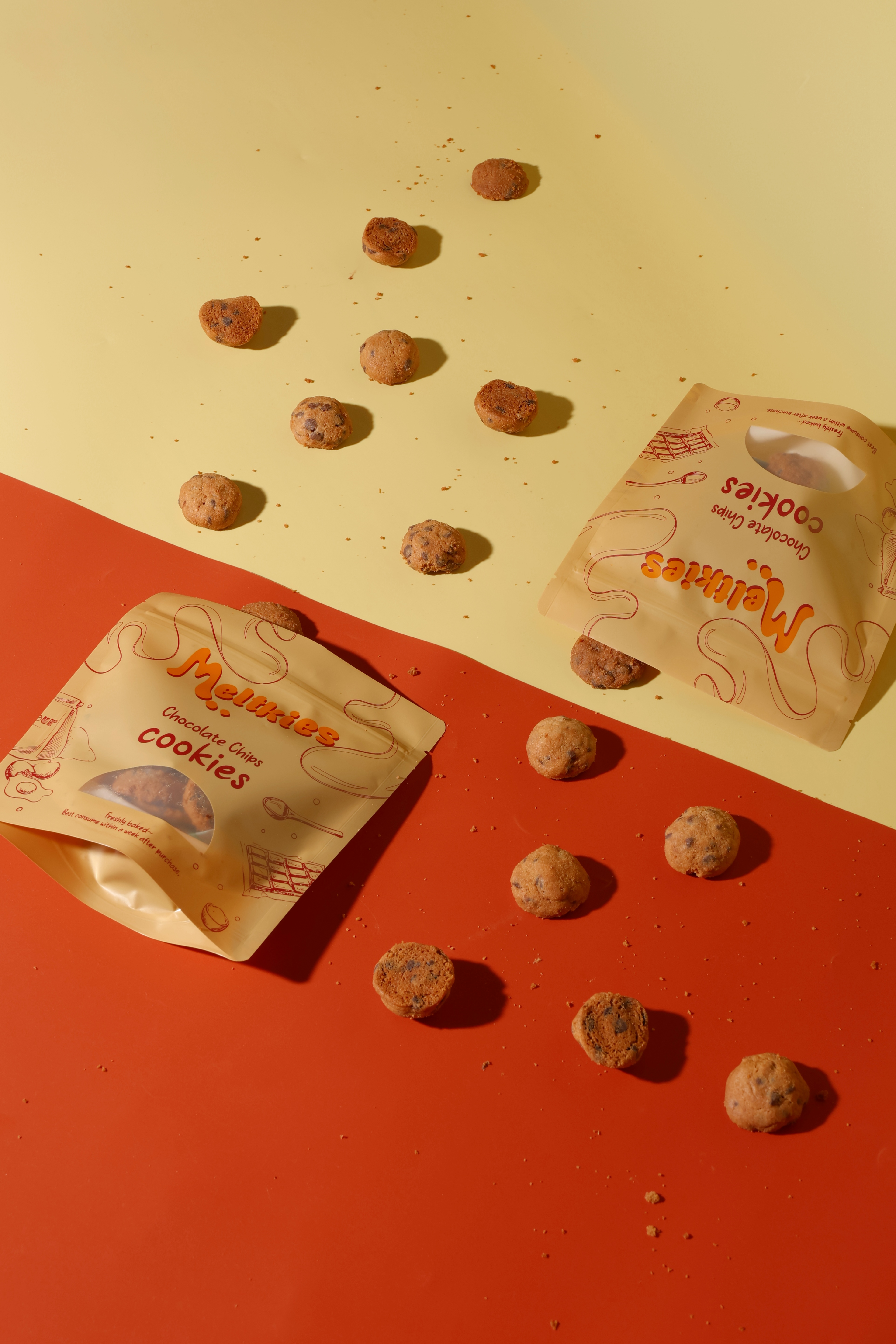 Two Melkkies Chocolate Chips Cookies packages with scattered small round cookies and crumbs on a yellow and red surface.