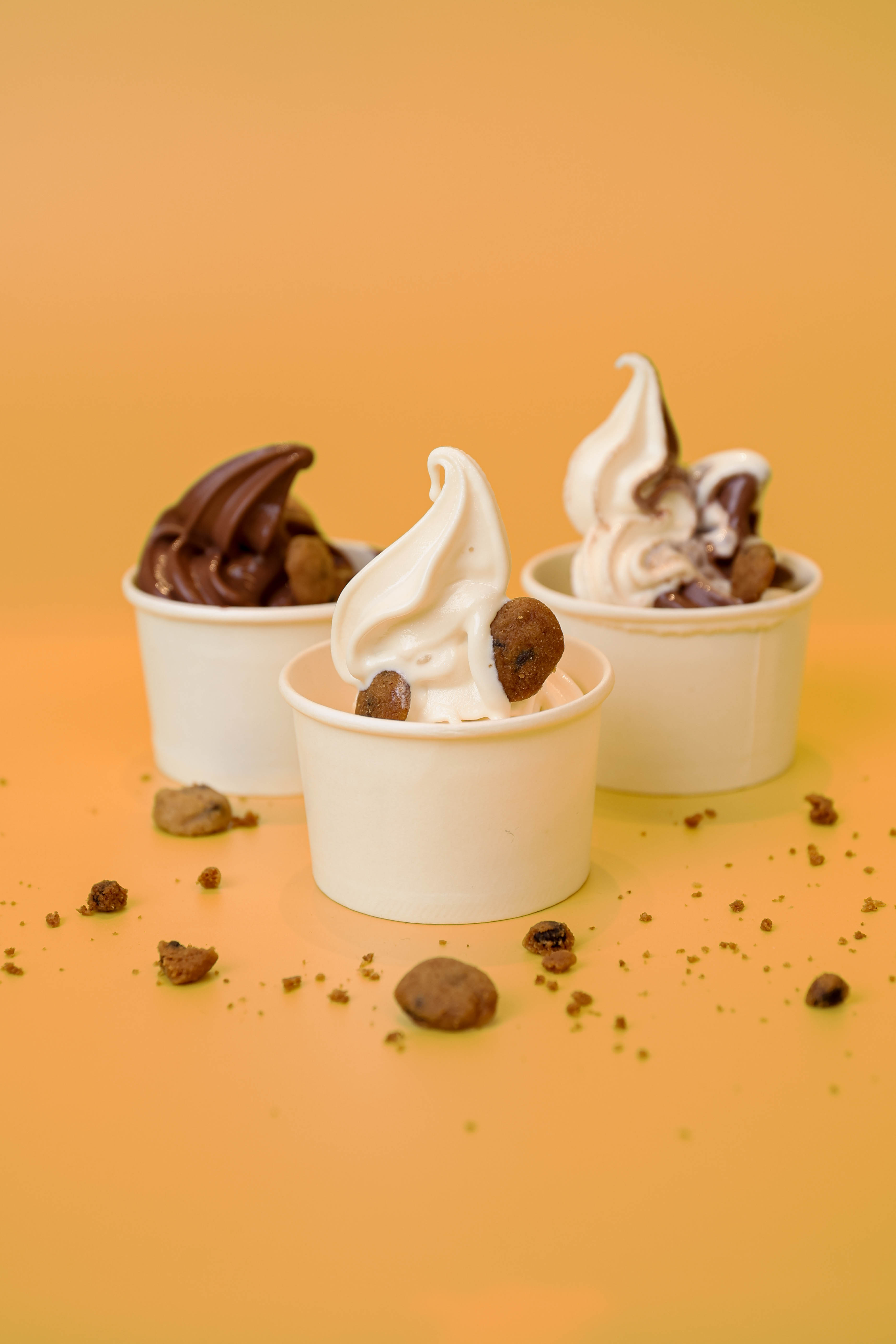 Three cups of soft serve ice cream with chocolate chip cookies on a yellow surface, featuring vanilla, chocolate, and a mixed swirl flavor.