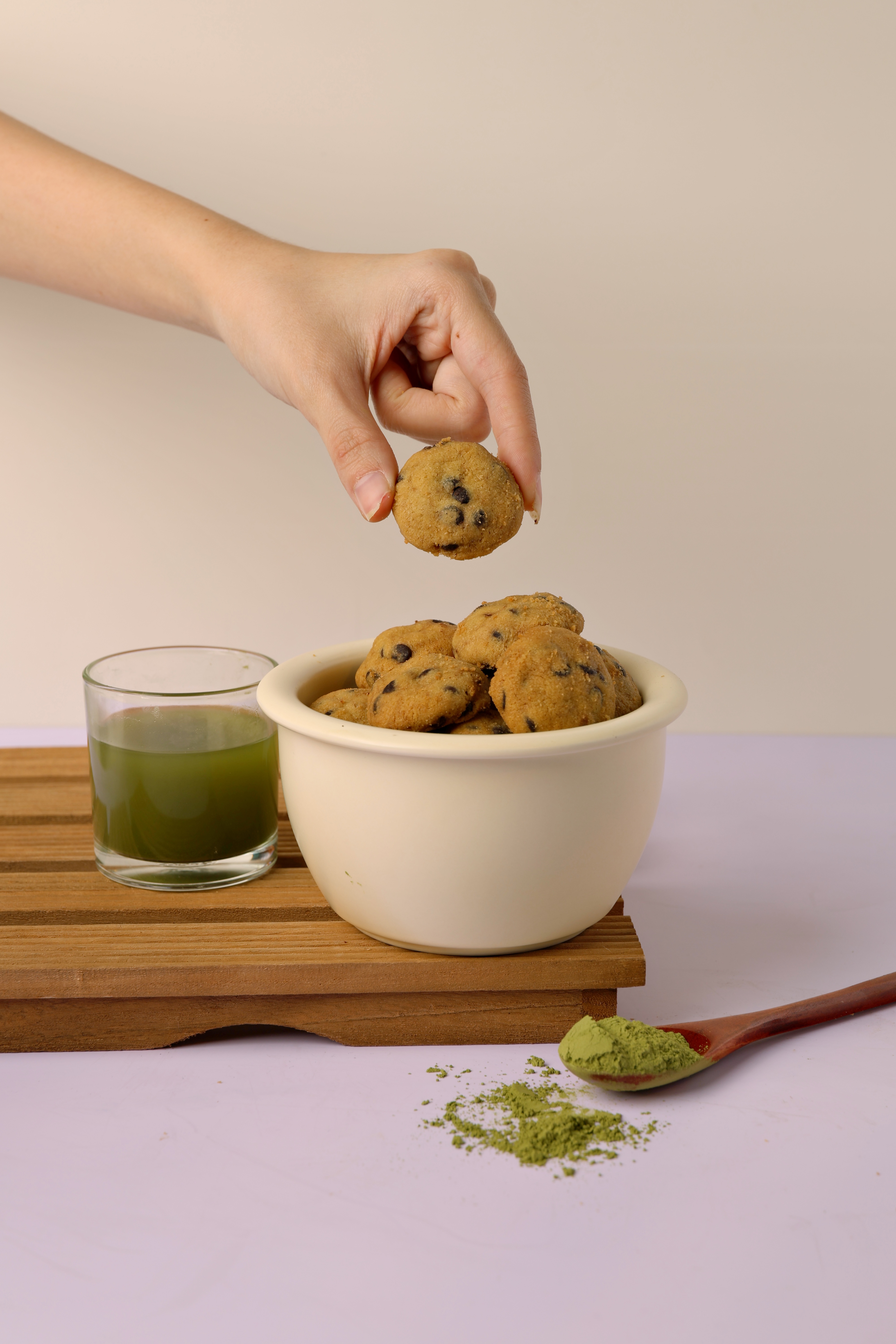 Hand picking up a chocolate chip cookie from a white bowl filled with cookies, next to a glass of green matcha tea and a wooden spoon with matcha powder.