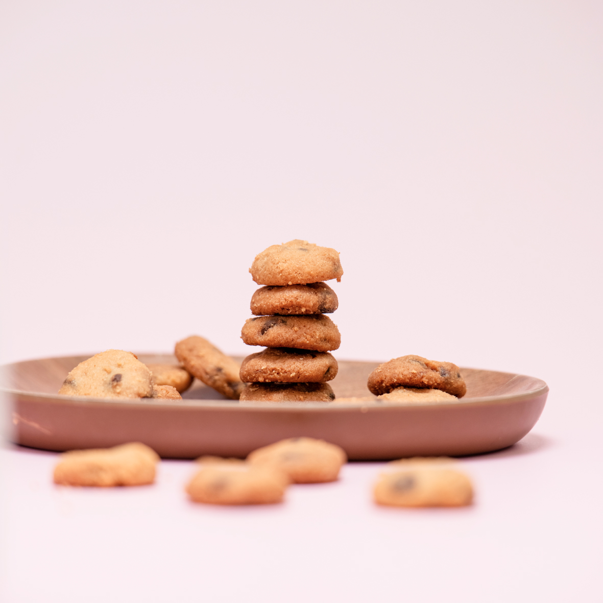 Stack of small chocolate chip cookies on a wooden plate with more cookies scattered around on a light pink background.