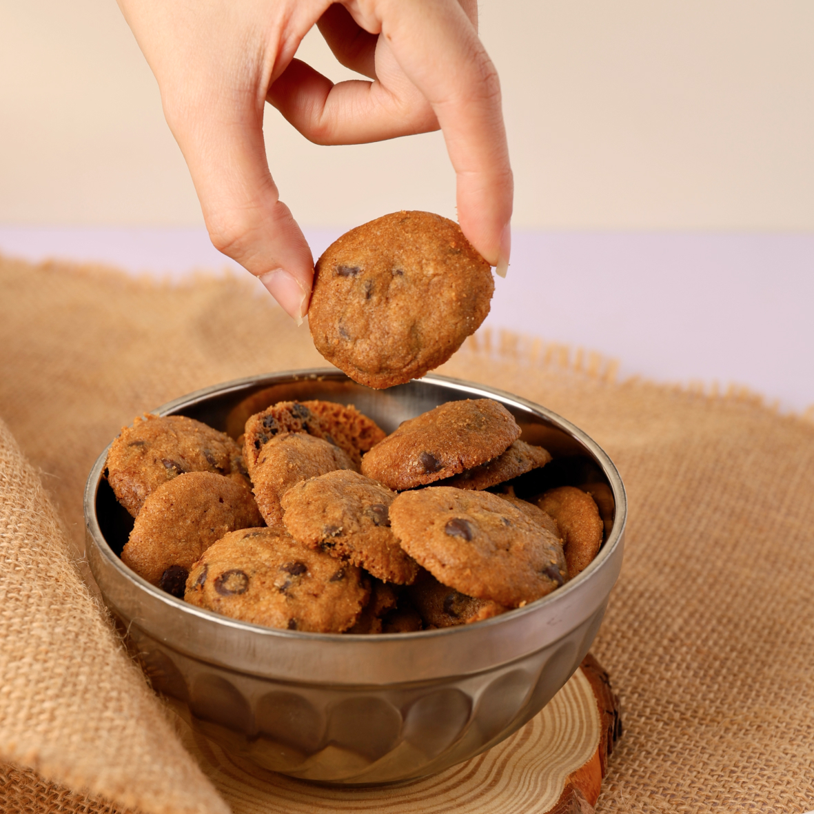 Hand picking a chocolate chip cookie from a metal bowl filled with cookies on burlap cloth.