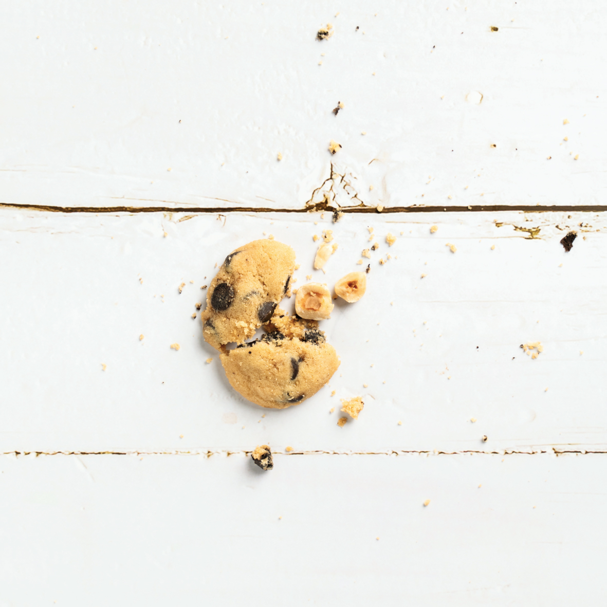 Broken chocolate chip cookie with two hazelnuts and crumbs on a white wooden surface.