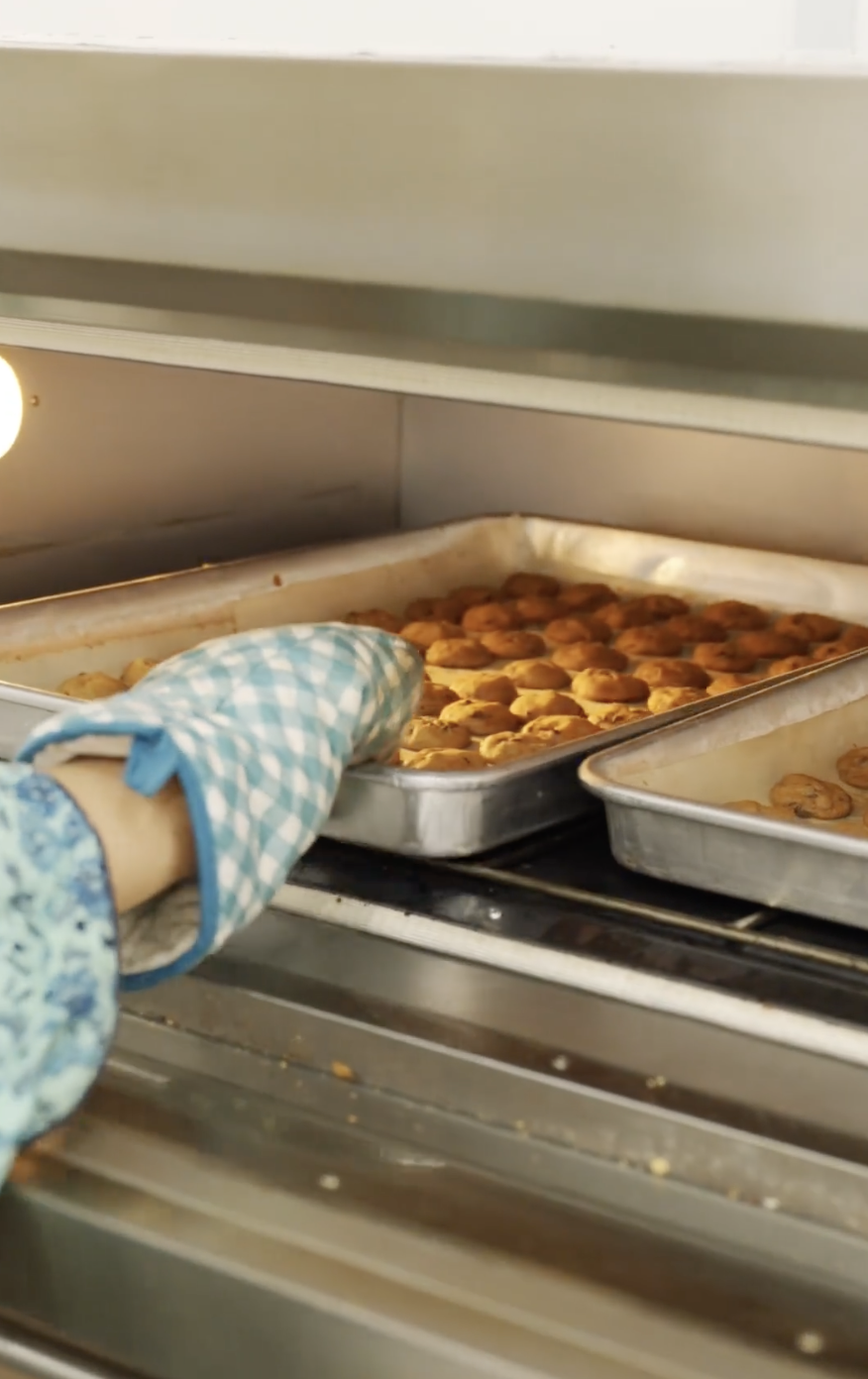 Person wearing a blue checkered oven mitt placing a tray of freshly baked cookies into an oven.