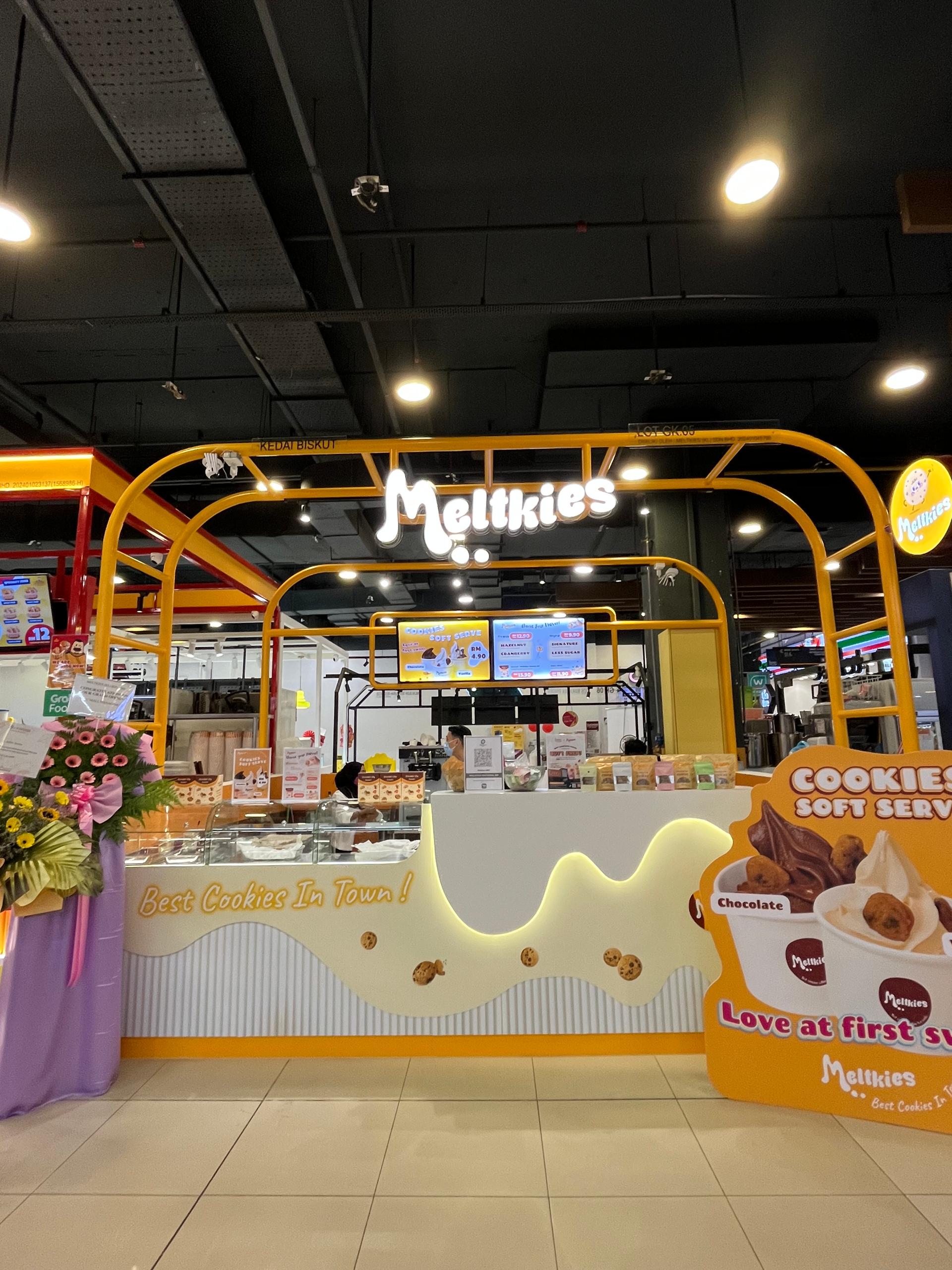 Melkties cookie shop kiosk with yellow frame, signage, flower stand, and display menus in a shopping mall.