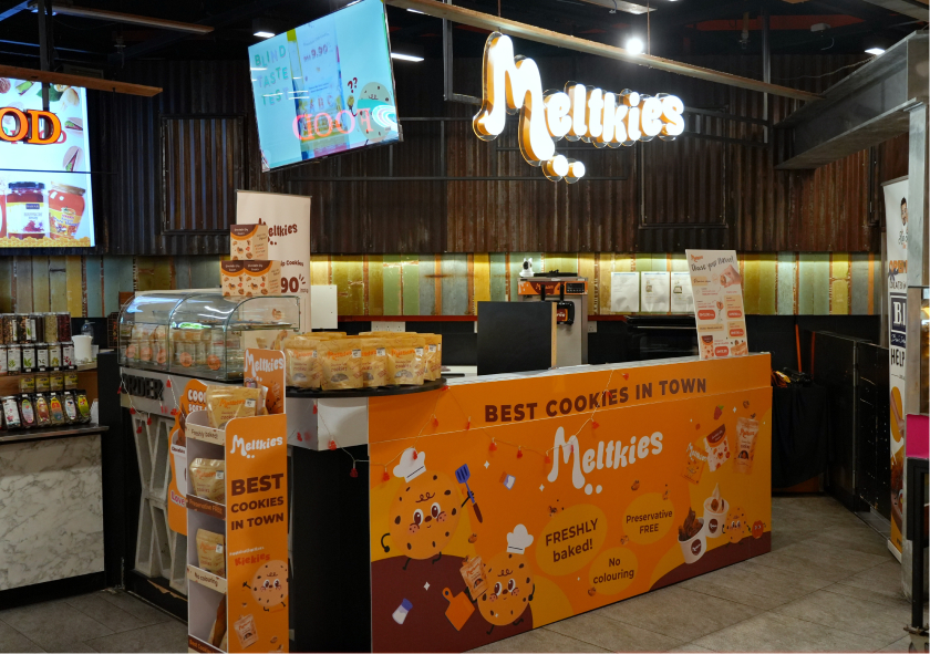 Meltkies cookie stand at a market with bright orange branding, featuring fresh-baked, preservative-free cookies.