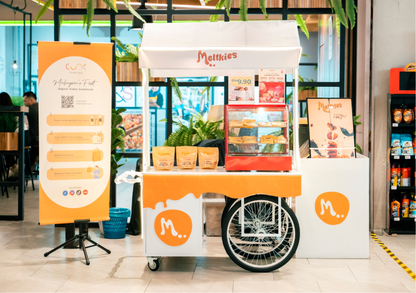 Melkties snack cart with packaged cookies and a display case of pastries inside a modern store with promotional banner on the left.