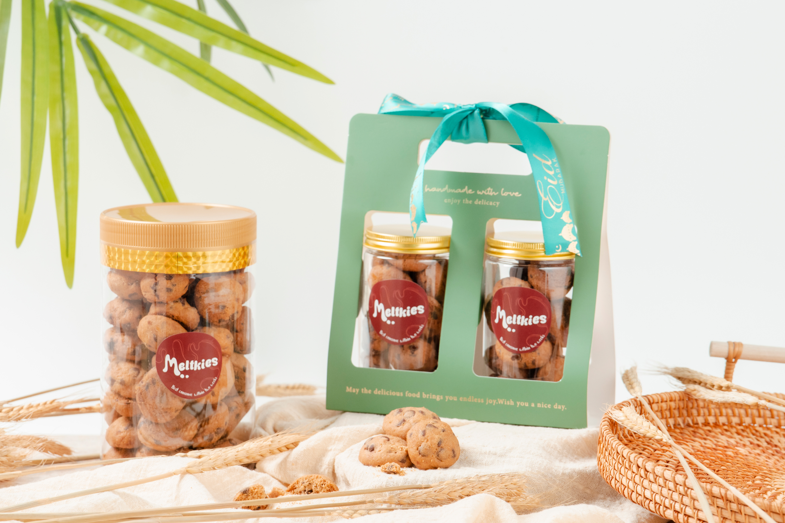 Two jars of Meltkies cookies with golden lids, one in green gift packaging with turquoise ribbon, surrounded by wheat stalks and a wicker basket on beige cloth.