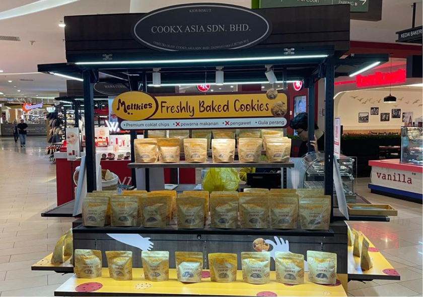 Display booth in a mall selling Meltkies freshly baked cookies with multiple packaged cookie bags arranged on shelves.