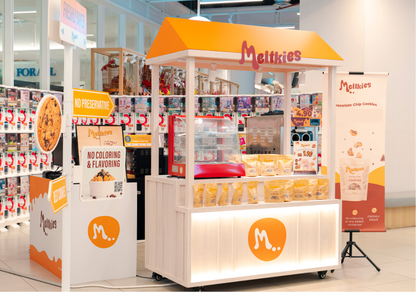 Melkies kiosk in a mall selling packaged chocolate chip cookies with signs stating no preservatives and no coloring or flavoring.