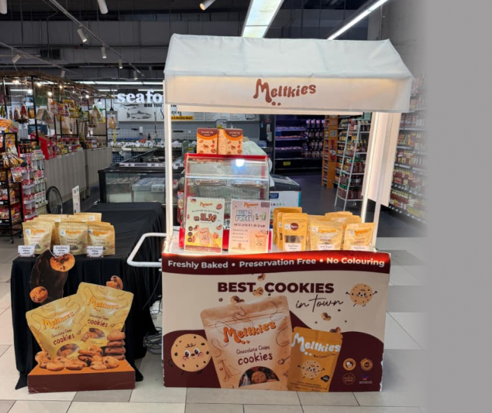 Melkkies cookie stand displaying bags of freshly baked, preservative-free chocolate chip cookies in a supermarket aisle.