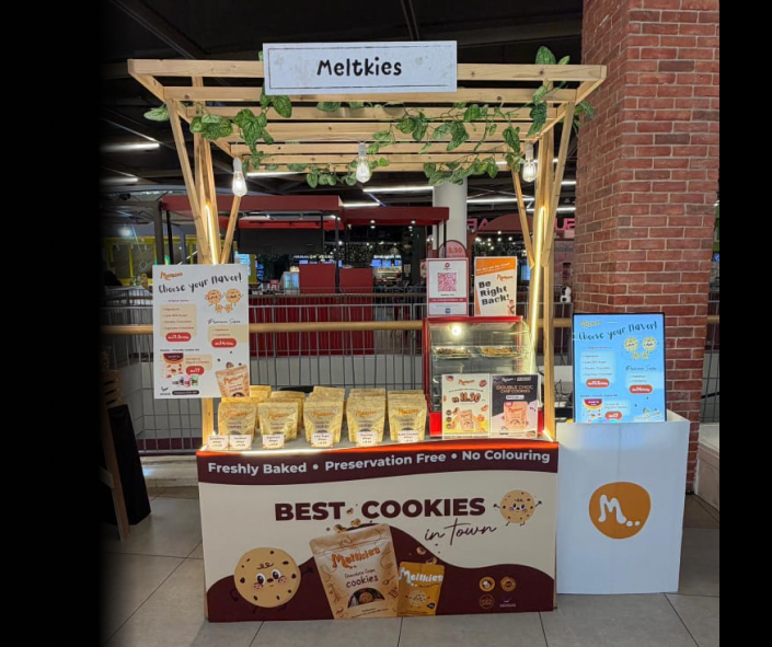 Meltkies cookie stand with wooden frame and green leaves decor, displaying various cookie packs with signs highlighting freshly baked, preservative-free, and no coloring.