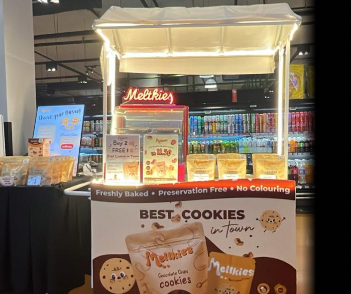 Retail display stall with illuminated sign 'Melkkies' offering freshly baked cookies labeled as preservation free and no coloring.