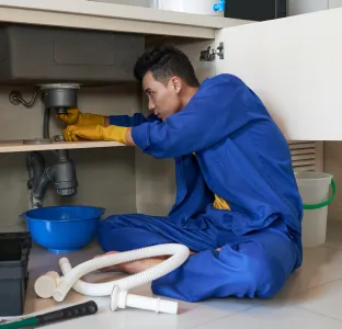 Plumber in blue coveralls and yellow gloves repairing pipes under a kitchen sink with tools and a blue basin nearby.