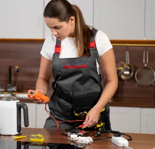 Woman in black overalls using a handheld device to test electrical wires on a kitchen counter.
