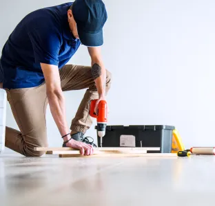 Man kneeling on the floor using an electric drill on a wooden plank with a toolbox and tools nearby.