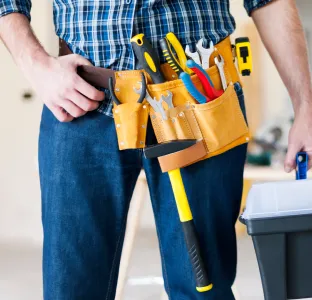 Person wearing a tool belt with various tools and holding a toolbox, dressed in a blue plaid shirt and jeans.