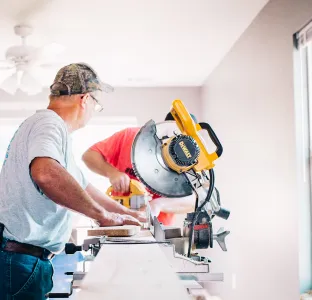 Two men using a yellow miter saw to cut wood indoors with natural light from a window.