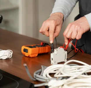 Person using a screwdriver to adjust a circuit breaker with a multimeter and cables on a wooden table.