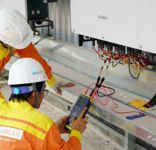 Two electricians in orange uniforms and white helmets testing electrical wiring connections with a handheld device.