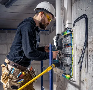 Electrician wearing a helmet and safety glasses working on an electrical panel.